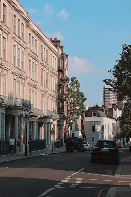 A view of a residential street in Lambeth with a row of elegant, multi-storey townhouses featuring large windows, decorative balconies, and stone facades. The street is lined with mature trees and parked cars along the curb. In the foreground, a bicycle lane is visible, with a few pedestrians walking on the sidewalk. In the background, modern high-rise buildings are visible against a blue sky with scattered clouds. The scene suggests a peaceful urban environment suitable for house removals and relocation services, with proximity to local amenities. The image provides a contextual setting for Man With a Van South Lambeth's house removal and furniture transport services, emphasizing the importance of careful packing and transportation logistics during home relocation.