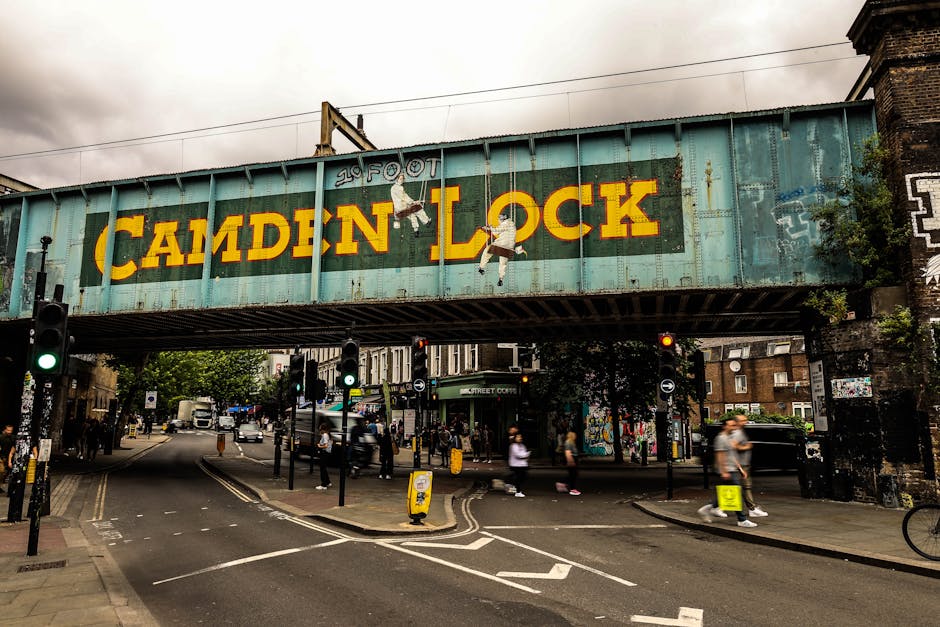 A large, weathered green railway bridge spans over a busy street in an urban area. The bridge features prominent yellow lettering spelling 'CAMDEN LOCK' with a warning about '20 FOOT' height and small illustrations of children hanging from the letters. Below the bridge, the street is filled with pedestrians walking on the sidewalks and crossing at the intersection, some of whom are slightly blurred to indicate movement. Several traffic lights are visible, with some showing green signals, and black bollards and yellow cone markers are placed along the road to manage vehicle flow. Buildings with various textures and graffiti surround the area, and cars, including a white van, are waiting or moving through the intersection. The scene is captured during daylight with overcast weather, creating a neutral, natural environment typical of a lively urban setting often visited during home relocations or furniture transport activities involving [COMPANY_NAME].