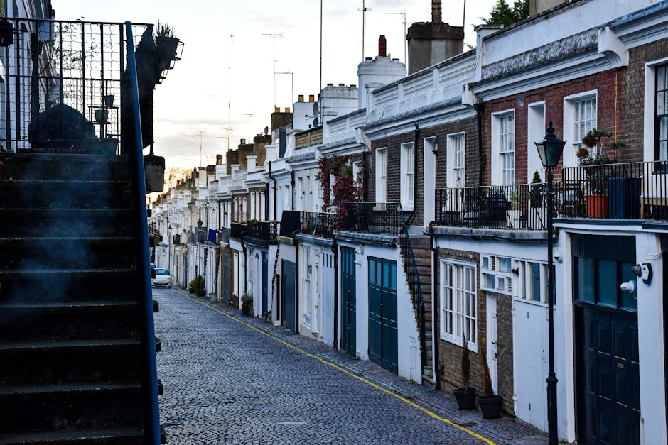 A view of a residential street in Lambeth with a row of elegant, multi-storey townhouses featuring large windows, decorative balconies, and stone facades. The street is lined with mature trees and parked cars along the curb. In the foreground, a bicycle lane is visible, with a few pedestrians walking on the sidewalk. In the background, modern high-rise buildings are visible against a blue sky with scattered clouds. The scene suggests a peaceful urban environment suitable for house removals and relocation services, with proximity to local amenities. The image provides a contextual setting for Man With a Van South Lambeth's house removal and furniture transport services, emphasizing the importance of careful packing and transportation logistics during home relocation.