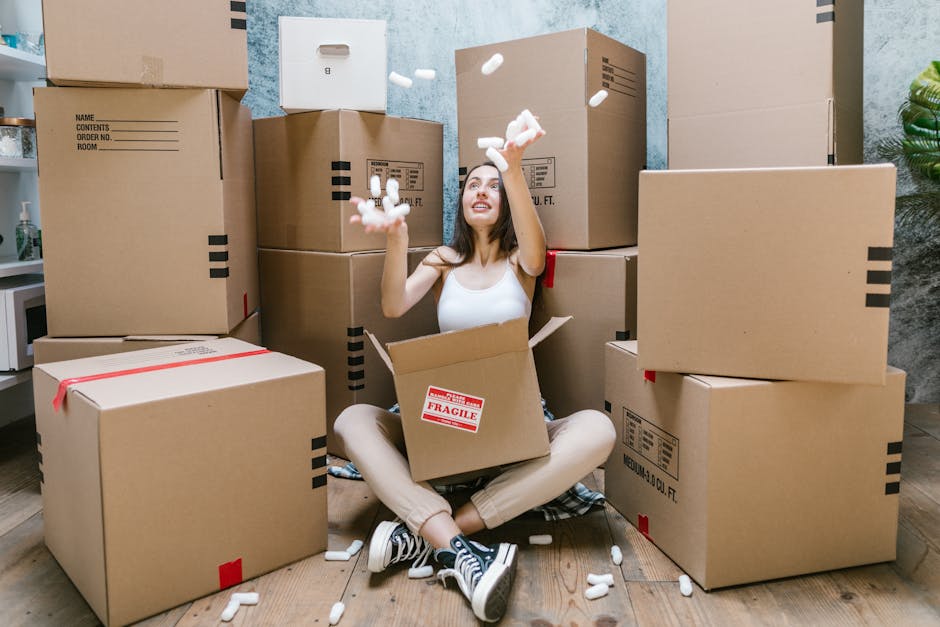 A young woman sitting cross-legged on a wooden floor inside an apartment, surrounded by numerous large cardboard moving boxes, some sealed with red tape and others open. She is wearing a white tank top, beige trousers, and black sneakers with white laces, and is smiling while tossing packing polystyrene foam pieces into the air. The boxes vary in size, with several stacked against the wall behind her, and labels indicating contents and fragile items. To the left, a small white shelf and part of a kitchen countertop are visible, while a decorative green plant is partly seen on the right. The setting is illuminated by natural or ambient indoor lighting, capturing a moment during home relocation and packing process, with [COMPANY_NAME] providing professional removals services for house and flat moves, as suggested by the scene of furniture transport and packing preparations.
