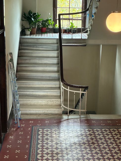 Inside a residential property, a staircase with marble steps and a dark wooden handrail curves upward from the ground floor, leading to an upper level. At the top of the stairs, a window with multiple panes allows natural light to illuminate the area, revealing potted plants in various sizes and colours placed on a checkered surface near the window sill. To the left of the staircase, a silver step ladder is positioned against the wall. The floor on the ground level features decorative ceramic tiles with a geometric pattern in shades of red, white, and black. A warm-toned wall-mounted light fixture emits a soft glow, and a grey wall partially surrounds the staircase. This scene depicts part of a home ready for furniture transport or packing as part of a house relocation, with the stairs and corridor area prepared for moving activities, typical of house removals services by Man With a Van South Lambeth.