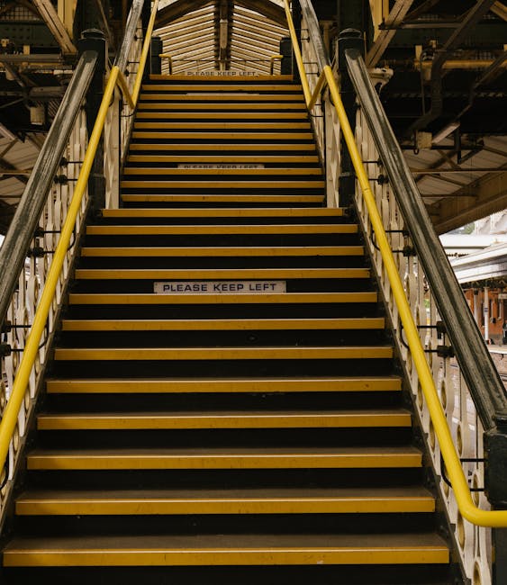 A steep metal staircase with yellow safety handrails on both sides leading upwards in an industrial setting, with visible structural beams and lighting overhead. The stairs feature yellow and black anti-slip strips on each step, and a sign on one of the steps reads 'PLEASE KEEP LEFT.' This stairway is located inside a building used for house removals or furniture transport, with some packaging materials and boxes possibly visible at the top or bottom areas, indicating a packing and moving environment. The scene suggests a careful and organized process of moving household items within or into a property, supported by professional removal services like Man With a Van South Lambeth.