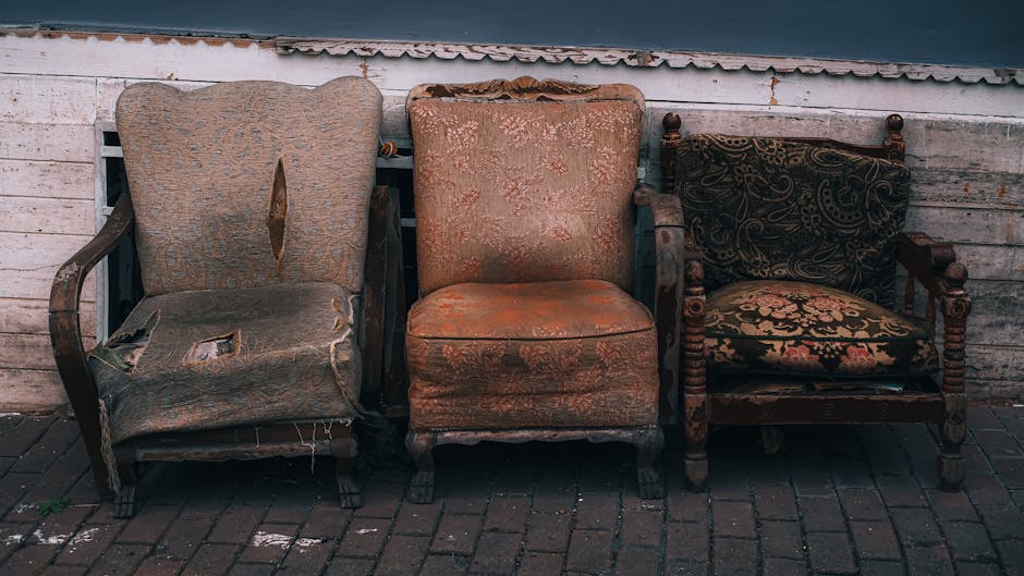 Three old, worn-out armchairs are positioned outside against a weathered white wooden wall on a paved surface. The first armchair on the left features damaged beige fabric upholstery with tears, exposing the padding and wooden frame beneath, and has wooden armrests showing signs of wear. The middle chair has faded, patterned orange upholstery with visible staining and slight fraying along the edges, and its wooden legs are partially visible. The third armchair on the right is crafted from dark polished wood with an elaborate carved design, including turned wooden detailing on the armrests and legs, and is upholstered in fabric with a black and beige floral pattern; it appears to be in slightly better condition than the others. The scene suggests a setting suitable for furniture removal or home relocation, with the chairs waiting to be loaded onto a moving vehicle, supported by the presence of a company such as Man With a Van South Lambeth that offers removals services for residential moves, furniture transport, and packing and moving logistics.