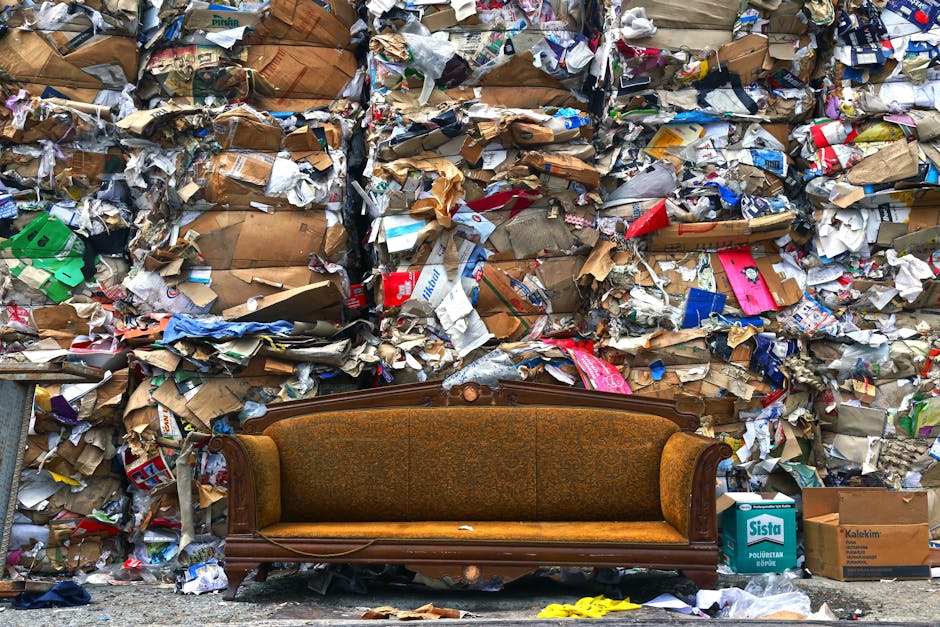 A vintage wooden sofa with an upholstered back and seat in a muted gold-brown fabric is positioned on a pavement in front of a large, overflowing pile of crumpled cardboard boxes, paper packaging, and plastic wrapping, all stacked against a wall. The surrounding area appears cluttered, indicating a waste collection or disposal scene. In the background, there are additional cardboard boxes and packaging materials, some labeled with brand names, along with a few scattered yellow objects on the ground. This setting reflects a disposal or recycling process associated with house removals or furniture transport, possibly related to the rules for disposing of old furniture as outlined by Lambeth Council, with the scene emphasizing the importance of proper waste management during home relocation projects. A professional removal service such as Man With a Van South Lambeth might assist in relocating or disposing of such items efficiently and responsibly.
