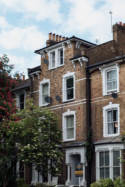 Photograph of a multi-storey brick residential building with white window frames and decorative white trim, situated on a street with trees and greenery in the foreground. The building has multiple chimneys on the roof, satellite dishes attached to the exterior walls, and a small shaded entranceway. The sky is partly cloudy with blue patches visible. This scene represents an urban residential environment suitable for home relocation or furniture transport, with the building’s exterior as the setting for potential house removals and moving services facilitated by companies like Man With a Van South Lambeth.