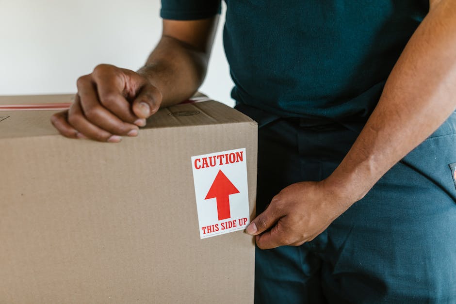 Close-up of a person wearing a dark green t-shirt and blue work trousers, positioning a large cardboard box with a white and red caution label indicating 'This Side Up' and an upward arrow; the individual is holding the box steady with their right hand, while their left hand is gripping the edge of the box. The scene takes place inside a residential property, with the person preparing to load or unload the box during a home relocation process. The background is neutral, emphasizing the packaging materials which are reinforced with brown packing tape. The image captures the careful handling and packing involved in furniture transport and box moving, illustrating the logistical aspect of professional removals, as offered by Man With a Van South Lambeth, supporting efficient packing and moving services to address delays and planning on the day of a house move.