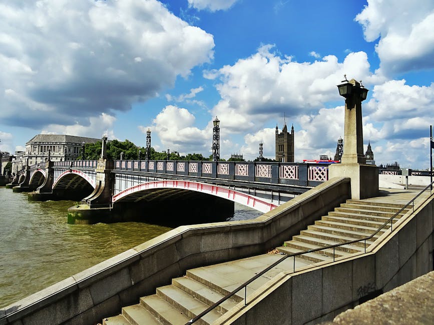 A view of Westminster Bridge spanning the River Thames in London, with the bridge's decorative ironwork painted in light colors and supported by stone arches. The river below has a slight greenish tint, and the sky above is partly cloudy with large, white fluffy clouds and patches of blue. On the right side of the image, a staircase made of light-colored stone leads down from a paved walkway, featuring metal handrails on both sides. Several tall, ornamental lamp posts line the bridge and walkway, and in the background, the historic clock tower of the Palace of Westminster, also known as Big Ben, is visible, along with surrounding buildings and towers characteristic of central London. The scene is well-lit with natural daylight, highlighting the architectural details and the busy urban setting, conveying a moment during a home relocation or moving process near the iconic London landmarks.