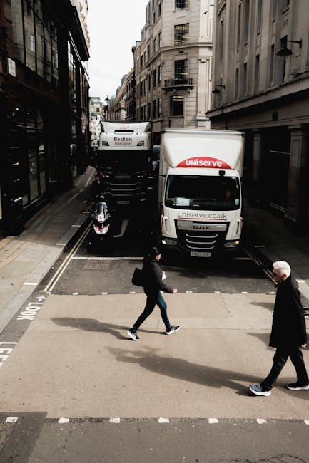The image depicts a narrow urban street scene during daylight, with multi-storey buildings on both sides. In the foreground, two pedestrians are crossing a designated pedestrian crossing on the road—one is a woman carrying a bag and dressed in dark clothing, while the other is an older man with white hair, dressed in dark trousers and a jacket. Behind them, a black scooter is parked close to the curb. Further back, two large white box trucks with logo and website branding are parked partially on the roadway, adjacent to a loading area. One truck bears the logo 'UNISERVE' on the front and side, with an open cargo area visible. The scene illustrates a typical street involved in a home relocation or furniture transport process, with the vehicles strategically positioned for loading or unloading, supporting the theme of professional removals by [COMPANY_NAME]. The lighting is natural, highlighting the urban environment during daytime.