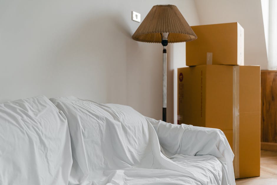 The image shows a section of a living room with a white sofa covered by a white sheet, positioned near a plain white wall. To the right of the sofa, there are three cardboard moving boxes stacked one on top of the other, with the largest at the bottom and the smallest at the top, all sealed with packing tape. A wooden piece of furniture, possibly a cabinet or sideboard, is partially visible behind the boxes. A tall, standing floor lamp with a conical brown lampshade is situated next to the boxes, casting light into the room. The scene appears to be part of a home relocation process, with packing materials and furniture ready for transport, highlighting the loading or packing stage of house removals. The environment is well-lit with natural or ambient light, and the setting suggests temporary storage or preparation for moving, consistent with professional removals activities by Man With a Van South Lambeth.