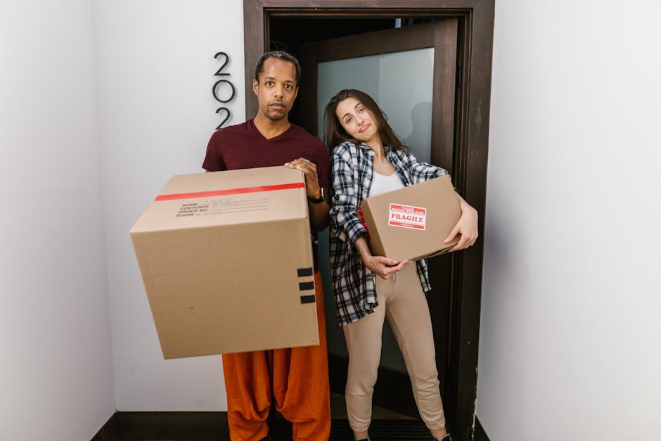 A man and a woman standing in a doorway inside a residential property, both holding cardboard boxes used for packing during the home relocation process. The man, dressed in a maroon t-shirt and orange pants, is holding a larger box with black shipping labels while the woman, wearing a checkered shirt over a white top and beige pants, is holding a smaller box with a red 'Fragile' sticker. The doorway features a dark wooden frame, and the wall next to it has the house number '202' mounted vertically. The interior lighting is bright, and nearby, moving supplies such as additional cardboard boxes and packing materials might be present, supporting a furniture transport and packing and moving process by [COMPANY_NAME], Man With a Van South Lambeth, as part of their professional removals service focused on house removals in Vauxhall SW8.