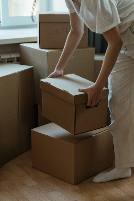 A person wearing light-colored clothing, including white trousers and socks, is seen inside a property, handling a small cardboard moving box during a home relocation. They are placing the box on top of a stack of larger cardboard boxes on a wooden floor near a window that allows natural light to illuminate the space. Additional packed boxes, some labeled, are arranged around them, indicating an organized packing process as part of a furniture transport or house removals project. The scene reflects careful handling and packing materials such as cardboard boxes used to safeguard belongings during a move, consistent with the services provided by Man With a Van South Lambeth. The environment appears tidy and bright, emphasizing the professional approach to packing and moving tasks involved in an internal or external house removal.